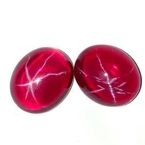 Puede incluir: Dos piedras preciosas de rubí estrella roja de forma ovalada con una estrella blanca en el centro.