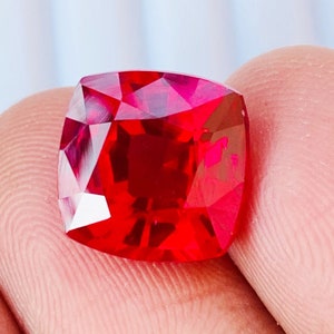 May include: A deep red gemstone with a cushion cut. The gemstone is faceted and has a high polish.