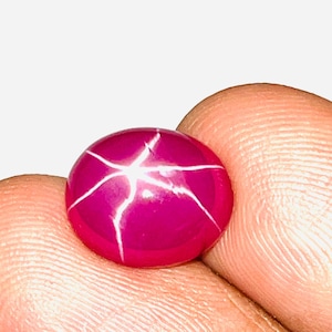 Może przedstawiać: Okrągły, polerowany kamień szlachetny rubinu z efektem gwiazdy. Klejnot ma głęboki różowy kolor z sześcioramiennym wzorem gwiazdy na powierzchni. Kamień szlachetny jest trzymany między dwoma palcami na białym tle.