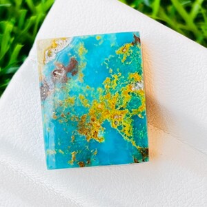 May include: A rectangular turquoise gemstone with a vibrant mix of blue, gold, and brown hues. The stone is set against a white surface, showcasing its unique colour patterns and textures. The gemstone's dimensions are not visible.