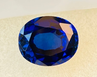 May include: A deep blue oval-shaped gemstone with facets, displayed in a white box.