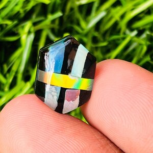 Op de afbeelding: Een zwarte, iriserende, zeshoekige cabochon edelsteen met een geometrisch patroon van wit, geel en roze.