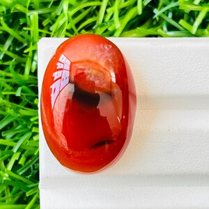以下が含まれることがあります： 鮮やかな赤色の、磨かれた楕円形の宝石。暗い渦巻き模様が特徴です。白い表面に置かれ、緑色の人工芝を背景に、印象的なコントラストを生み出しています。この宝石は、おそらくジュエリーや装飾目的で使用されます。