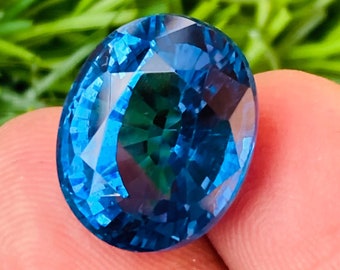 May include: A vibrant blue oval-shaped gemstone with a faceted cut. The gemstone is held in a hand, with a green grassy background.