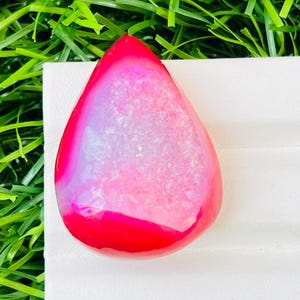 Può includere: Una gemma a forma di goccia con una vibrante sfumatura di colore. La gemma passa da un bordo rosso intenso a un centro rosa e bianco tenue. La pietra è posta su una superficie bianca ed erba verde.