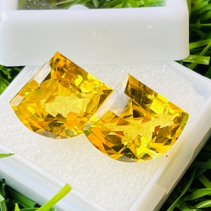 May include: Two faceted, yellow gemstones in a white plastic case, set against a backdrop of green grass. The gemstones have a fan-like shape and a brilliant, transparent appearance, reflecting light.