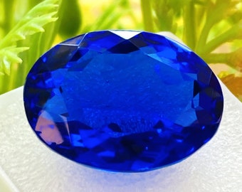 May include: A large, oval-shaped, deep blue gemstone with a faceted cut. The gemstone is sitting on a white, textured surface.