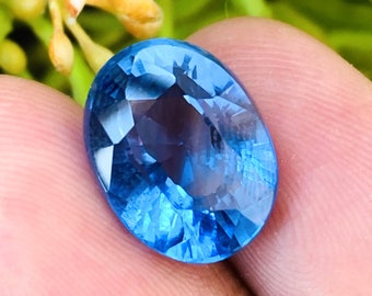 May include: A vibrant blue oval-shaped gemstone with a faceted cut. The gemstone is held in a hand, showcasing its brilliance and clarity.