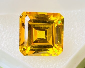 May include: A bright yellow gemstone with a square emerald cut. The gemstone is set in a white box.