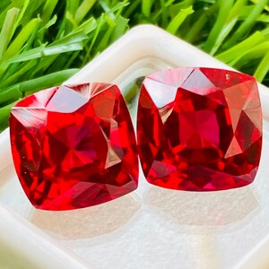 May include: Two bright red square-shaped gemstones in a white display box. The gemstones are cut with many facets and have a high shine.