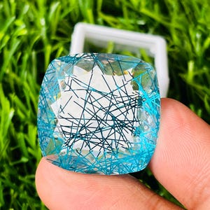 May include: A cushion-cut blue rutilated quartz gemstone featuring a transparent light blue body with dark, needle-like inclusions crisscrossing throughout the interior. The faceted stone displays a geometric pattern on its surface.