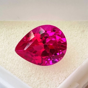 May include: A vibrant pink pear-shaped gemstone, with a faceted cut, is displayed in a white box.