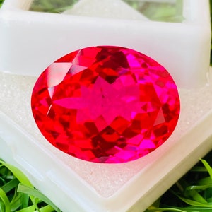 Peut inclure: Une grande pierre précieuse rouge vif de forme ovale avec de nombreuses facettes. La pierre précieuse est posée dans une boîte blanche sur un lit d'herbe verte.