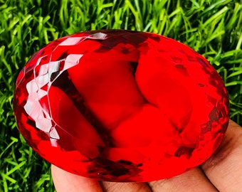May include: A large, faceted, red crystal in the shape of an oval. The crystal is being held in a hand, with green grass in the background.