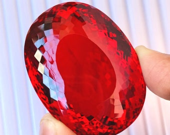 May include: A large, oval-shaped, red gemstone with a faceted cut. The gemstone is held in a hand.