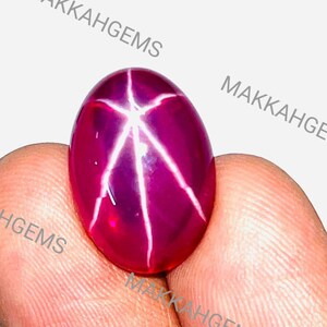 Peut inclure: Une pierre précieuse polie, de forme ovale, rouge foncé, avec un motif en étoile à six rayons. L'étoile est blanche et ressort sur le fond rouge intense. La pierre est tenue entre les doigts, avec le texte "MAKKAHGEMS" visible.