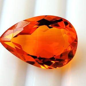 May include: A faceted, teardrop-shaped gemstone in a bright orange colour. The gem's surface reflects light, displaying its intricate cuts and polished facets. The gemstone is set against a white background, highlighting its colour and clarity. Suitable for jewellery.
