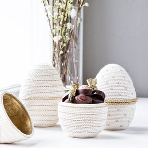 Può includere: Tre contenitori a forma di uovo di Pasqua in crochet bianco con accenti dorati. Un contenitore è aperto e pieno di caramelle al cioccolato.