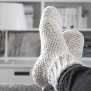 May include: A pair of white crocheted socks with a ribbed cuff. The socks are being worn on someone's feet and are resting on a grey surface.