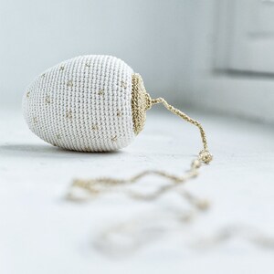 Può includere: Un ornamento a forma di uovo in crochet bianco con dettagli dorati e una catena dorata.