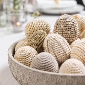 Może przedstawiać: Miska wypełniona szydełkowymi jajkami wielkanocnymi w różnych odcieniach beżu i złota. Jajka są ułożone w dekoracyjny wzór, tworząc świąteczny i uroczy wygląd.