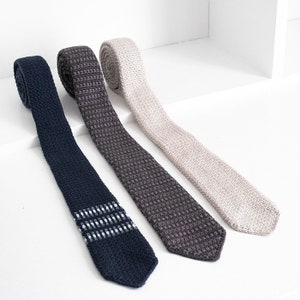 May include: Three knitted neckties in different colors: navy blue with white stripes, dark gray, and light beige. The ties are made of a textured knit fabric.