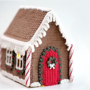 Op de afbeelding: Een gehaakt peperkoekhuis met een rode deur, witte versiering en snoepstokaccenten. Het huis heeft een krans op de deur en een raam met een lichtje erin.