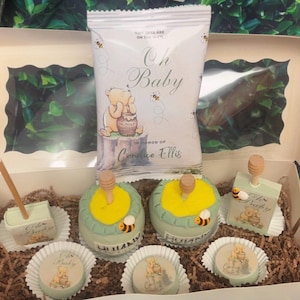 May include: A baby shower gift box featuring Winnie the Pooh themed treats. The box includes a bag with "Oh Baby" text and illustrations, along with decorated cakes and chocolates. The treats are decorated in yellow, green, and brown.
