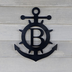May include: Black nautical-themed wall decor featuring an anchor design with a ship's wheel and the letter "B" in the center. The anchor is a classic design, and the piece is mounted on a light gray wooden background.