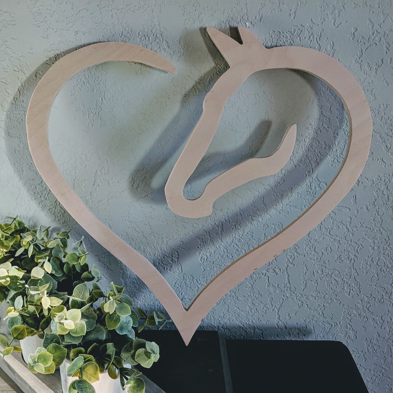 May include: A wooden cutout of a horse's head inside a heart shape. The cutout is painted in a light wood color and is mounted on a light blue wall.