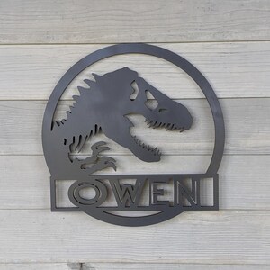 Op de afbeelding: Zwarte metalen wanddecoratie met een dinosaurus silhouet in een cirkel met de naam "OWEN" eronder.