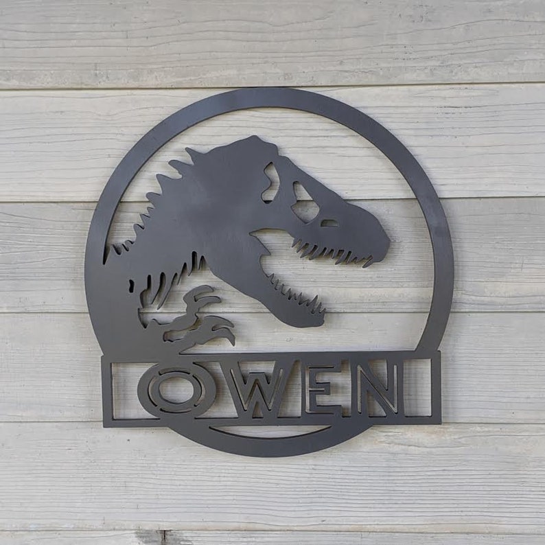 Op de afbeelding: Zwarte metalen wanddecoratie met een dinosaurus silhouet in een cirkel met de naam "OWEN" eronder.