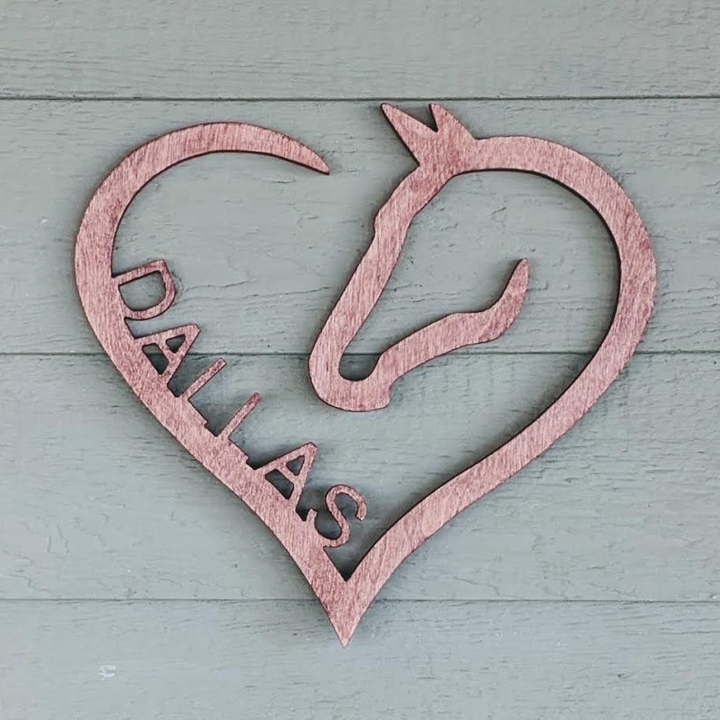 May include: Wooden heart-shaped sign with a horse head silhouette and the name "DALLAS" cut out inside. The sign is stained a light brown color.