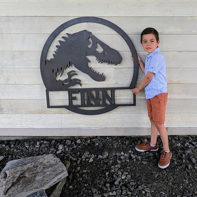 Op de afbeelding: Een zwart metalen dinosaurus silhouet met de naam "FINN" eronder. Een jonge jongen staat naast het dinosaurus silhouet.