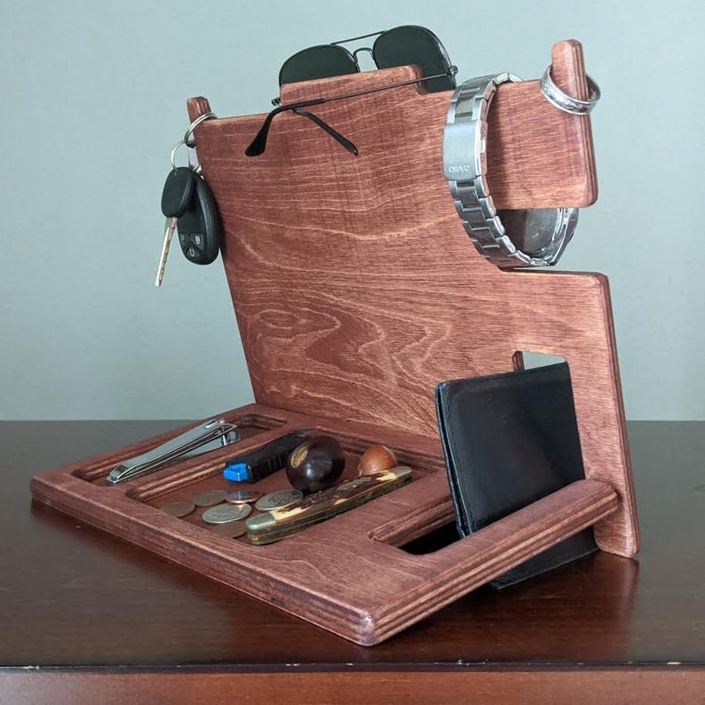 May include: A wooden organizer with slots for keys, a watch, sunglasses, and a wallet. It has a tray for loose items and a small compartment for a pocket knife. The organizer is stained a dark reddish-brown color.