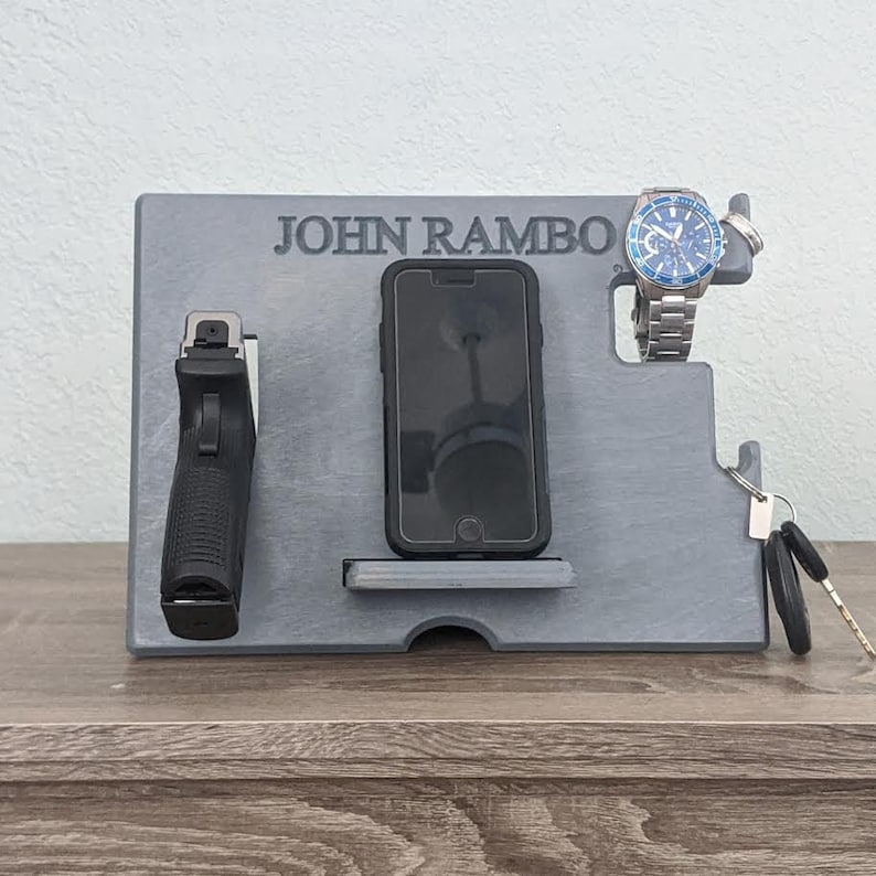 K&ouml;nnte beinhalten: Ein grauer Holzorganizer mit dem eingravierten Namen "JOHN RAMBO". Er hat F&auml;cher f&uuml;r ein Telefon, eine Uhr, Schl&uuml;ssel und eine Waffe.