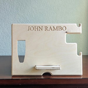 K&ouml;nnte beinhalten: Ein heller Holz-Telefonst&auml;nder mit einer Aussparung f&uuml;r ein Telefon und einem kleinen Regal zum Aufladen. Der St&auml;nder tr&auml;gt den Namen "JOHN RAMBO", der oben eingraviert ist.