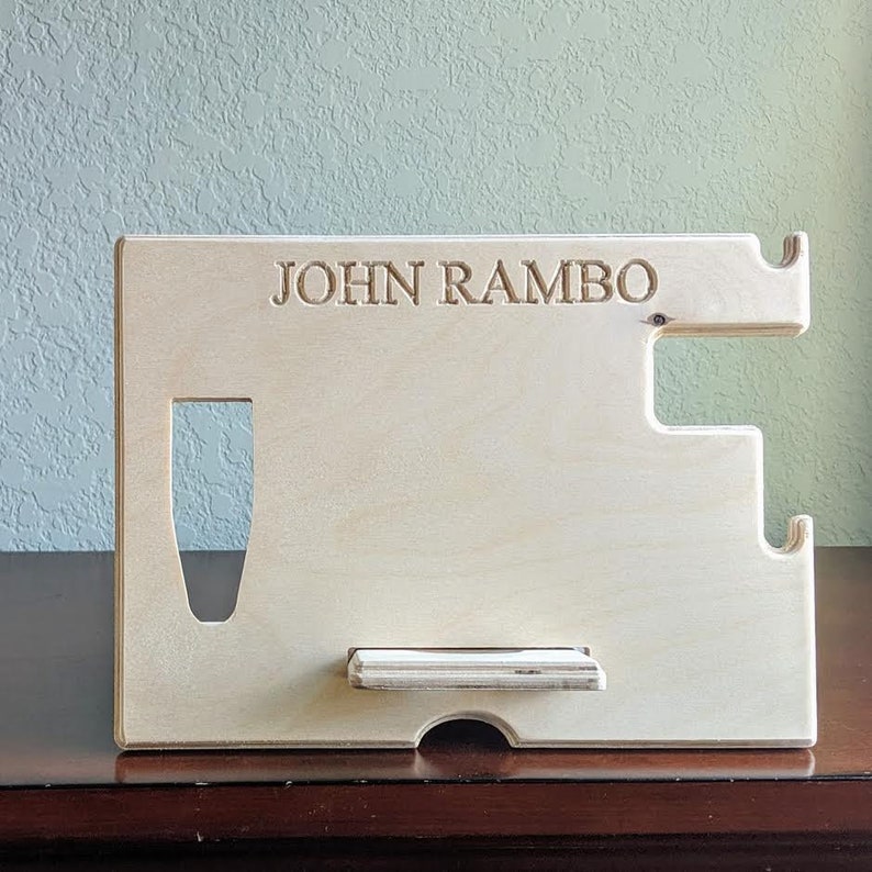 K&ouml;nnte beinhalten: Ein heller Holz-Telefonst&auml;nder mit einer Aussparung f&uuml;r ein Telefon und einem kleinen Regal zum Aufladen. Der St&auml;nder tr&auml;gt den Namen "JOHN RAMBO", der oben eingraviert ist.