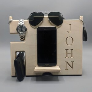 May include: A wooden organizer with the name "JOHN" engraved on the front. It has slots for a watch, phone, sunglasses, and keys.