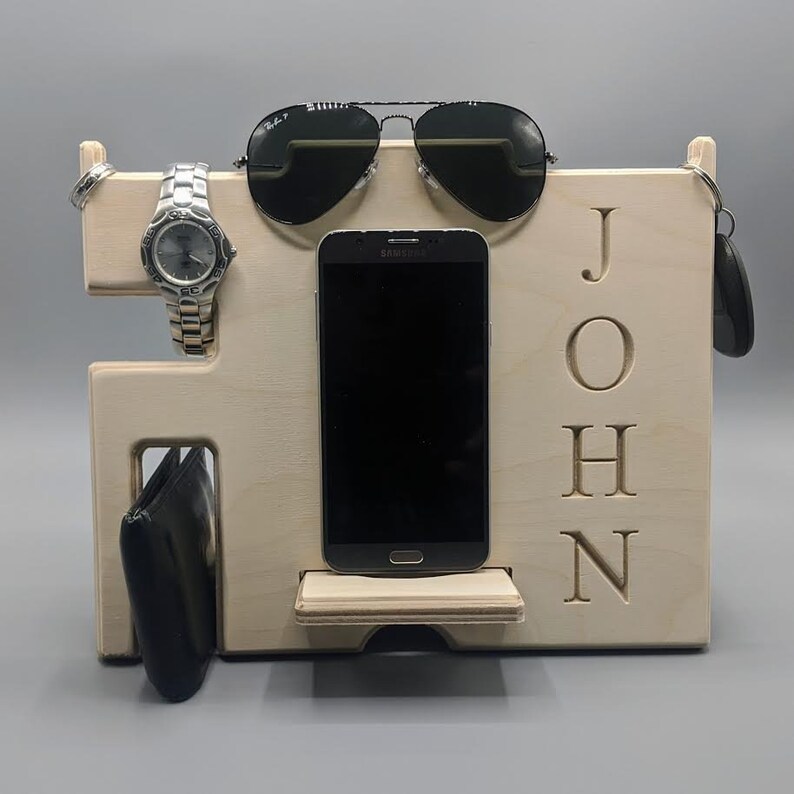 May include: A wooden organizer with the name "JOHN" engraved on the front. It has slots for a watch, phone, sunglasses, and keys.