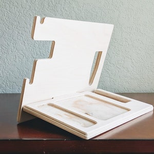 K&ouml;nnte beinhalten: Ein wei&szlig;er Holz-Telefonst&auml;nder mit mehreren Schlitzen zum Aufladen von Ger&auml;ten. Der St&auml;nder hat einen erh&ouml;hten R&uuml;cken und eine flache Oberfl&auml;che zum Ablegen eines Telefons.