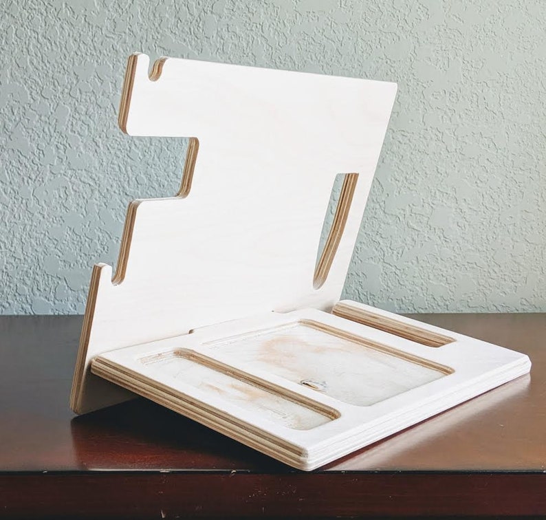 K&ouml;nnte beinhalten: Ein wei&szlig;er Holz-Telefonst&auml;nder mit mehreren Schlitzen zum Aufladen von Ger&auml;ten. Der St&auml;nder hat einen erh&ouml;hten R&uuml;cken und eine flache Oberfl&auml;che zum Ablegen eines Telefons.