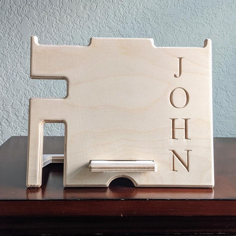 May include: A wooden phone stand with a cutout for a phone and a small shelf. The stand is personalized with the name "JOHN" in a vertical orientation.