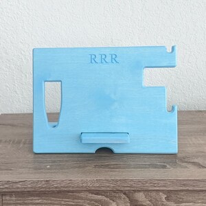 K&ouml;nnte beinhalten: Ein hellblauer, h&ouml;lzerner Telefonst&auml;nder mit einem Ausschnitt f&uuml;r ein Telefon und einem kleinen Regal zum Aufladen. Der St&auml;nder hat die Buchstaben "RRR" auf der Vorderseite eingraviert.