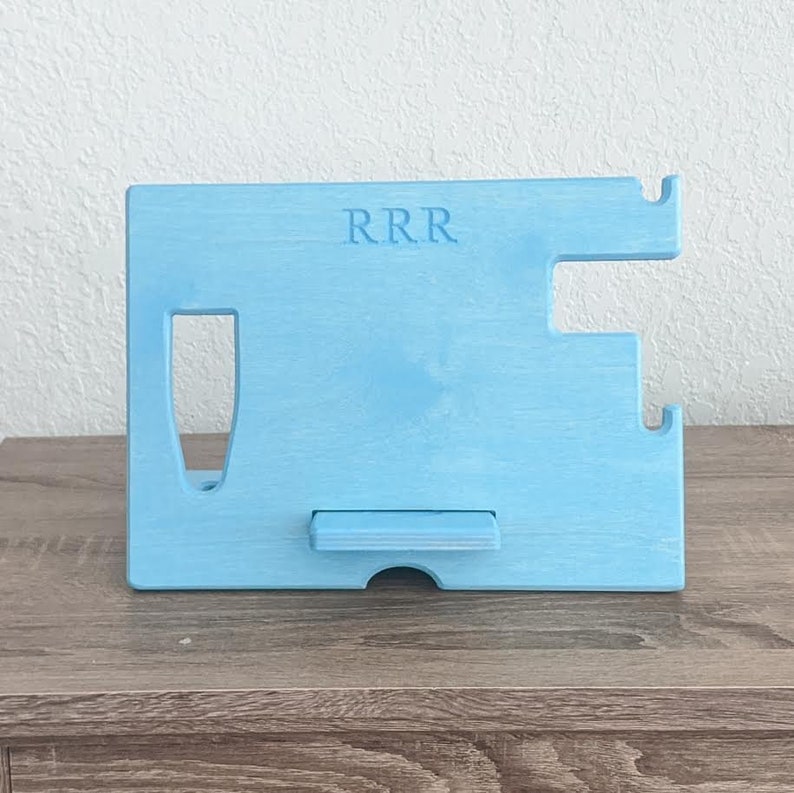 K&ouml;nnte beinhalten: Ein hellblauer, h&ouml;lzerner Telefonst&auml;nder mit einem Ausschnitt f&uuml;r ein Telefon und einem kleinen Regal zum Aufladen. Der St&auml;nder hat die Buchstaben "RRR" auf der Vorderseite eingraviert.