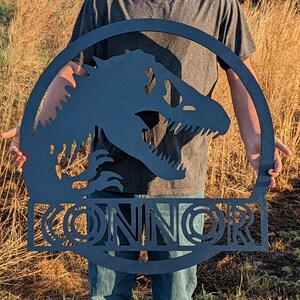 Op de afbeelding: Zwarte metalen silhouet van een dinosaurus met de naam "CONNOR" eronder. Het silhouet heeft de vorm van een cirkel.