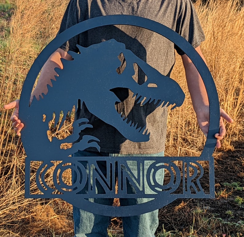 Op de afbeelding: Zwarte metalen silhouet van een dinosaurus met de naam "CONNOR" eronder. Het silhouet heeft de vorm van een cirkel.