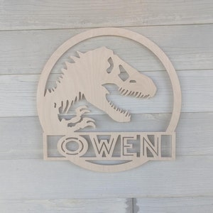 Op de afbeelding: Houten dinosaurus silhouet met de naam "OWEN" eronder uitgesneden. De dinosaurus bevindt zich in een cirkel met een kleinere cirkel binnen de grotere cirkel.