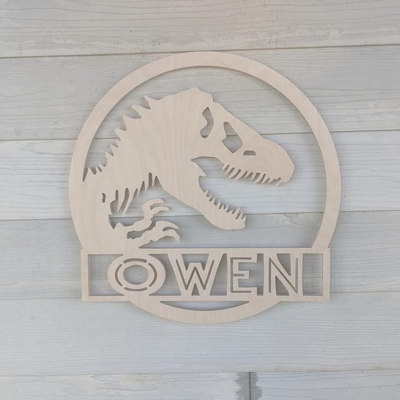 Op de afbeelding: Houten dinosaurus silhouet met de naam "OWEN" eronder uitgesneden. De dinosaurus bevindt zich in een cirkel met een kleinere cirkel binnen de grotere cirkel.