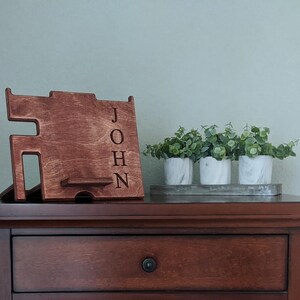 May include: A wooden phone stand with the name "JOHN" engraved on the side. The stand is sitting on a wooden table with a drawer and three small potted plants.